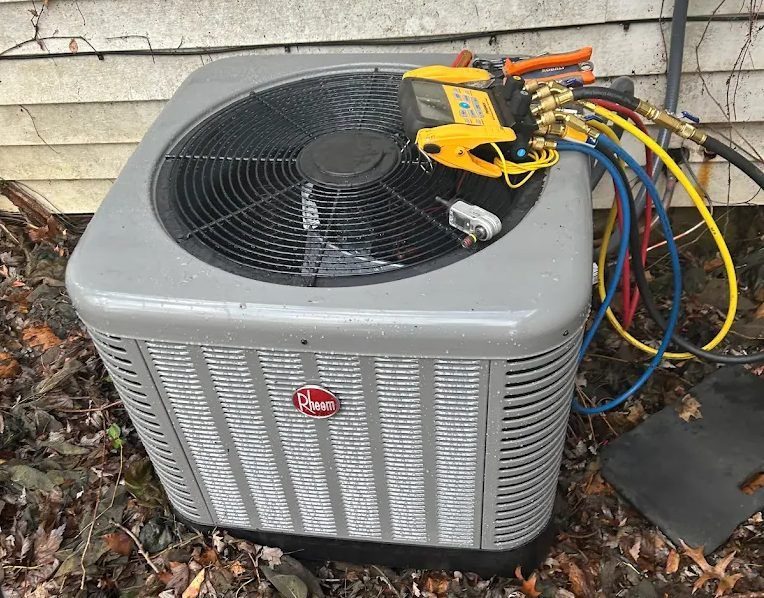 Outdoor Rheem air conditioning condenser with a yellow refrigerant manifold gauge and colored hoses attached for servicing outside a house.