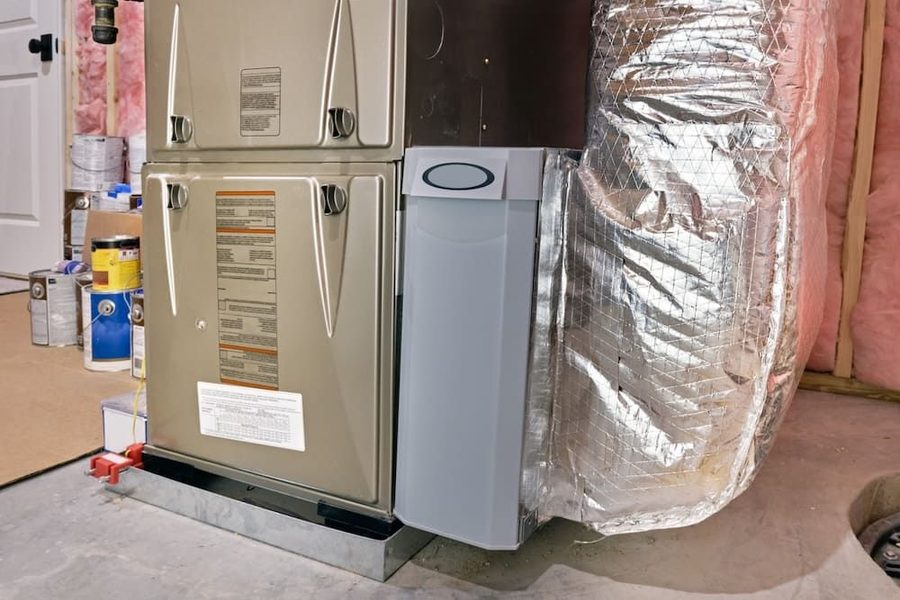 Beige furnace in an unfinished basement with foil-wrapped ductwork beside it and pink insulation in the wall. Furnace unit with a gray side panel and insulated metal ducting in a utility room/basement setting.