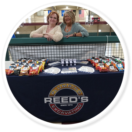 Two people stand on an athletic court in front of a table with promotional Reed's swag